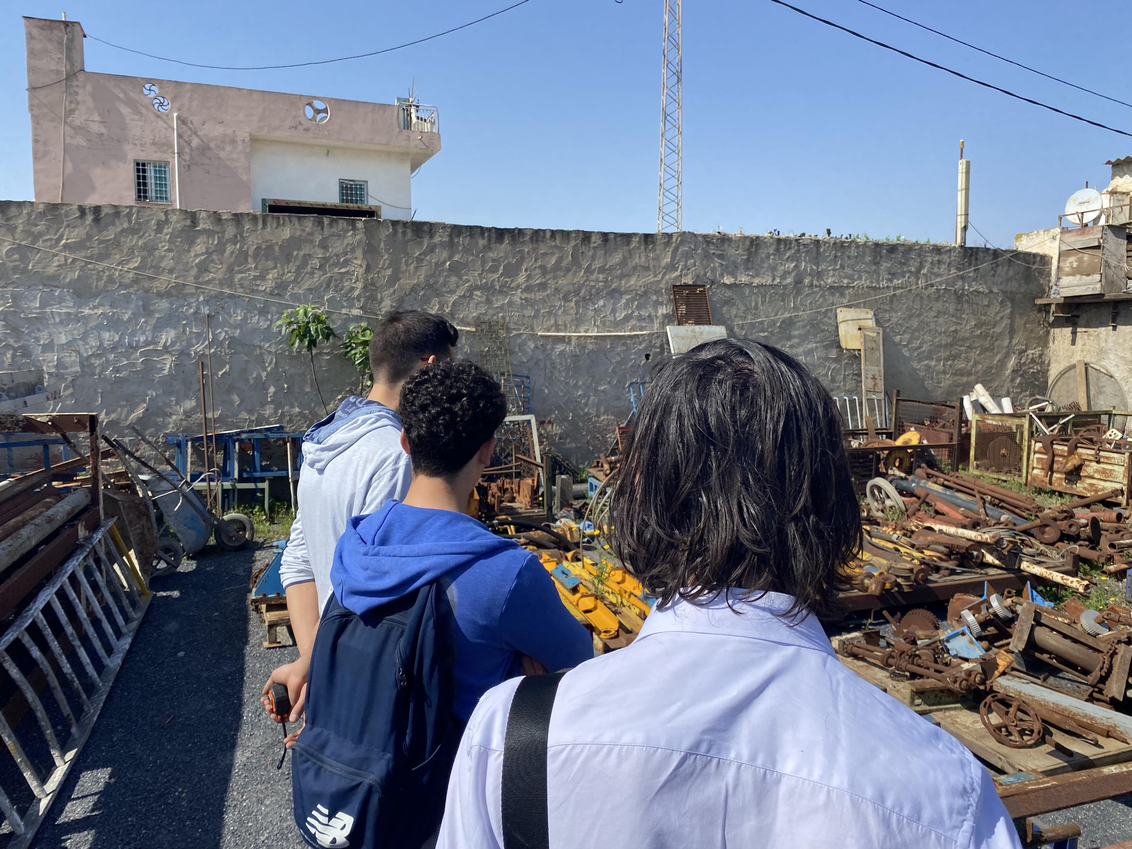 Three people facing rows of salvaged metal and vehicle parts in a scrapyard