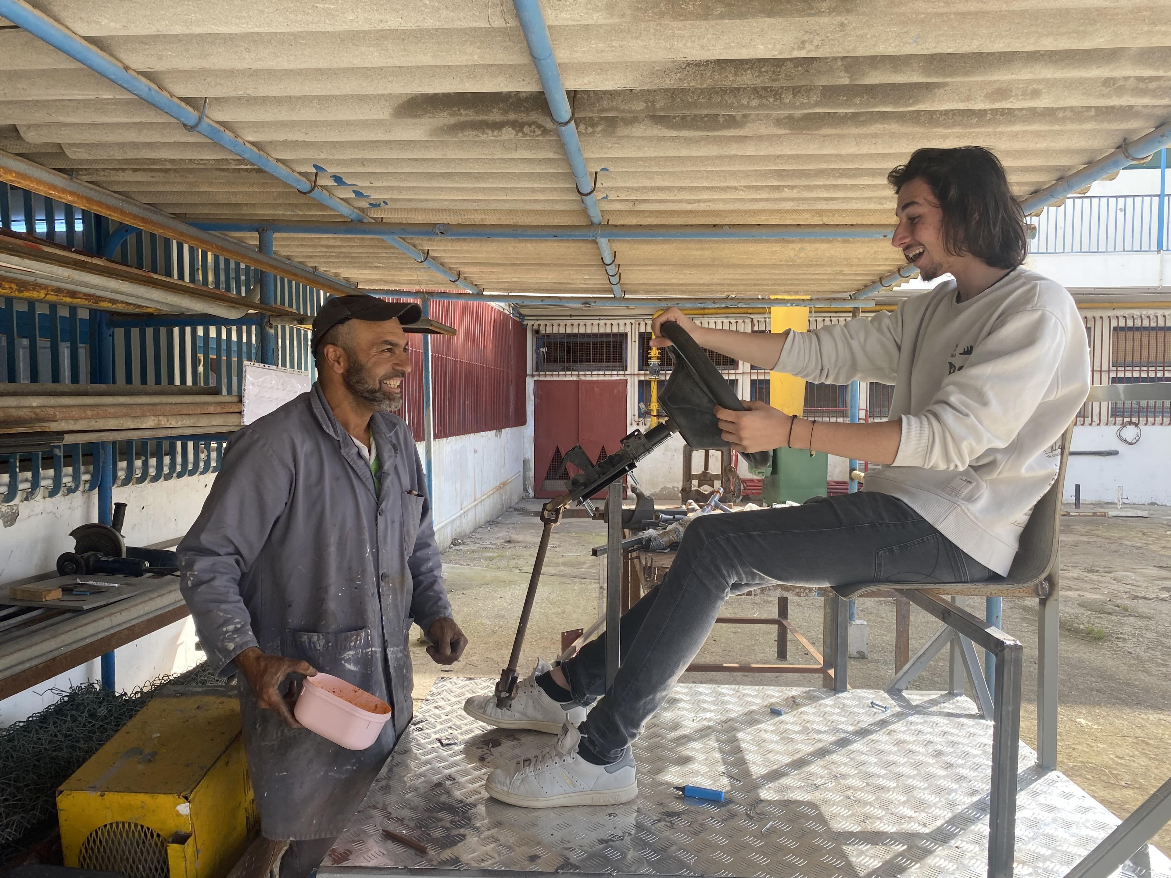 Two people beside the steering wheel and seat of the unfinished vehicle in a workshop