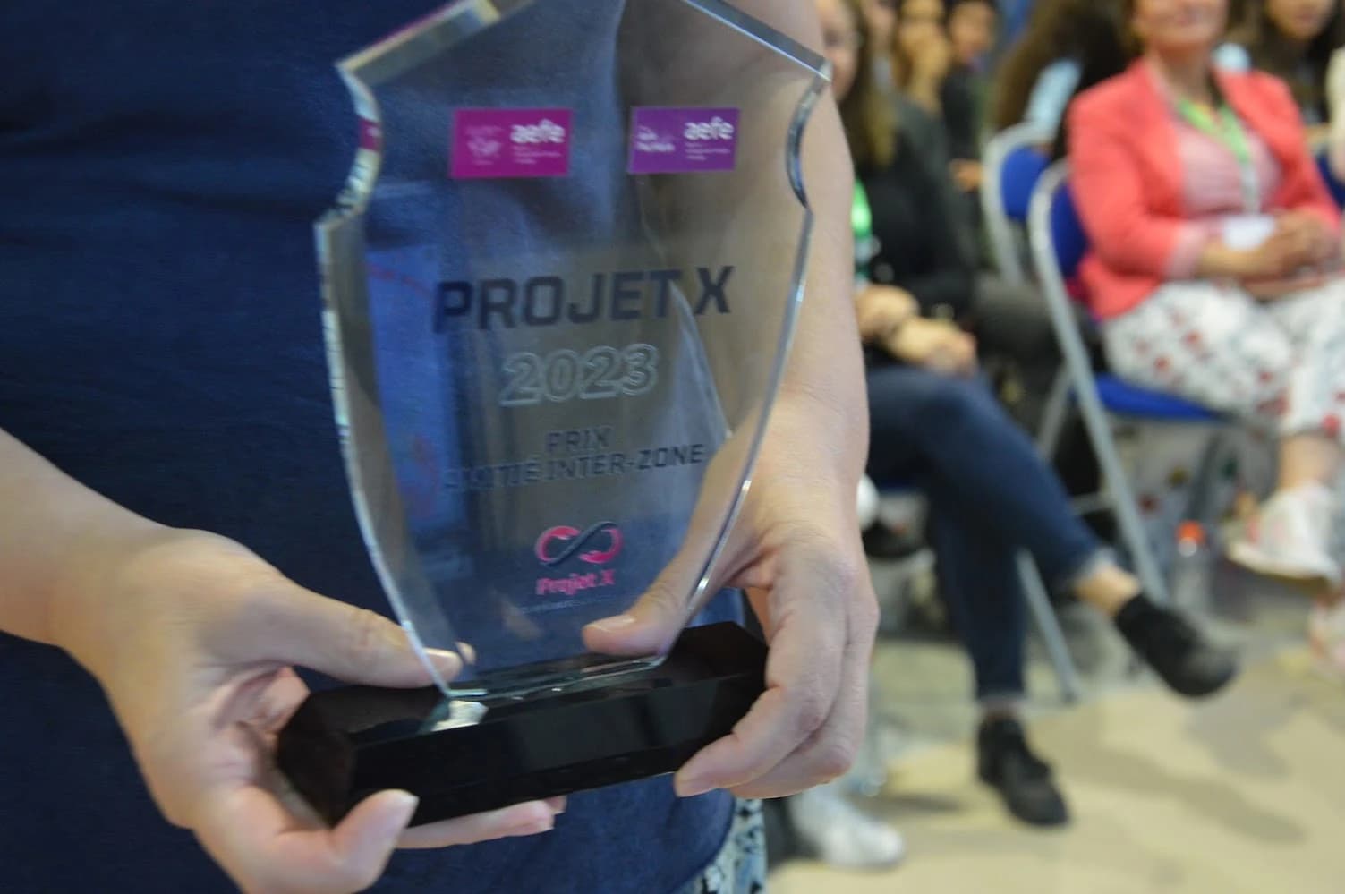 Un trophée en verre gravé « Projet X 2023 » et « Prix lycéen inter-zone », tenu à deux mains